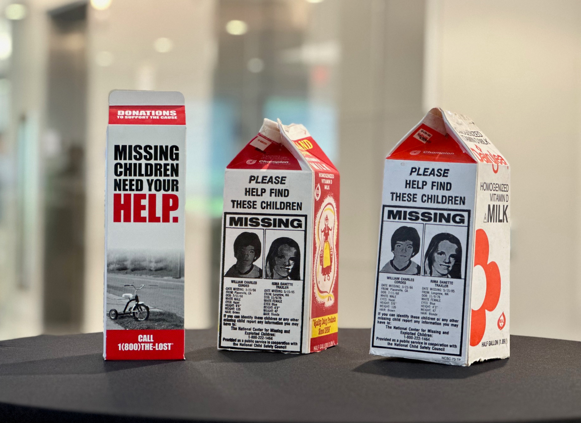 three red, white, and black cardboard milk cartons lined up on a table, each featuring missing children posters